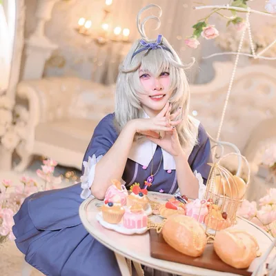 คอสเพลเยอร์ในชุด Robin จาก Honkai: Star Rail นั่งยิ้มที่โต๊ะน้ำชาซึ่งเต็มไปด้วยขนมอบและดอกไม้ในห้องสีขาวหรูหรา