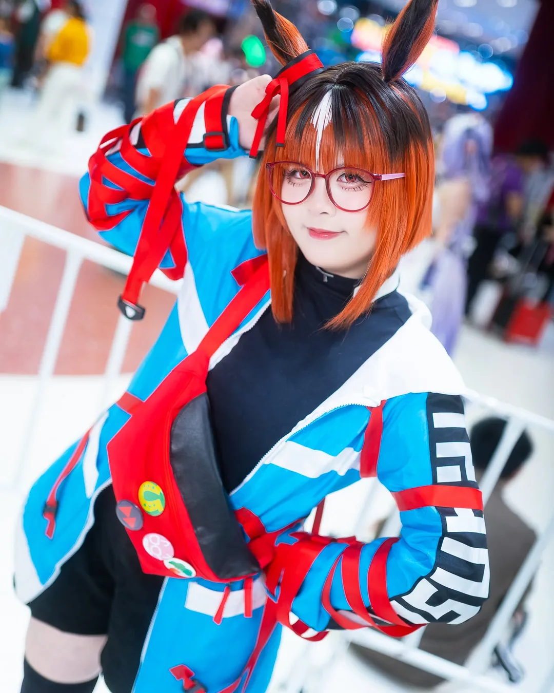 A cosplayer dressed as Dantsu Flame from Uma Musume poses with red glasses and animal ears at an indoor convention.