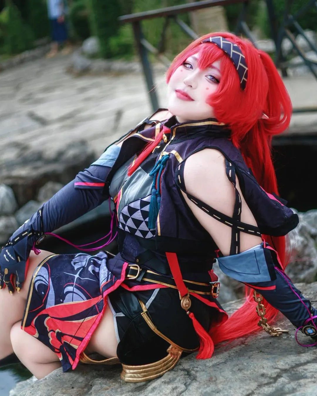 A cosplayer dressed as Yinlin with long red hair sits and smiles on a rock beside a garden pond.