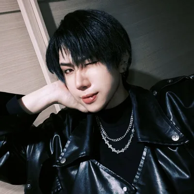 คนผมสั้นสีดำกำลังขยิบตาพร้อมเอามือเท้าศีรษะ สวมแจ็กเก็ตหนังสีดำและสร้อยคอโซ่เงินอยู่ภายในอาคาร