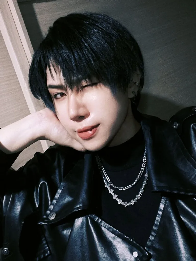 คนผมสั้นสีดำกำลังขยิบตาพร้อมเอามือเท้าศีรษะ สวมแจ็กเก็ตหนังสีดำและสร้อยคอโซ่เงินอยู่ภายในอาคาร