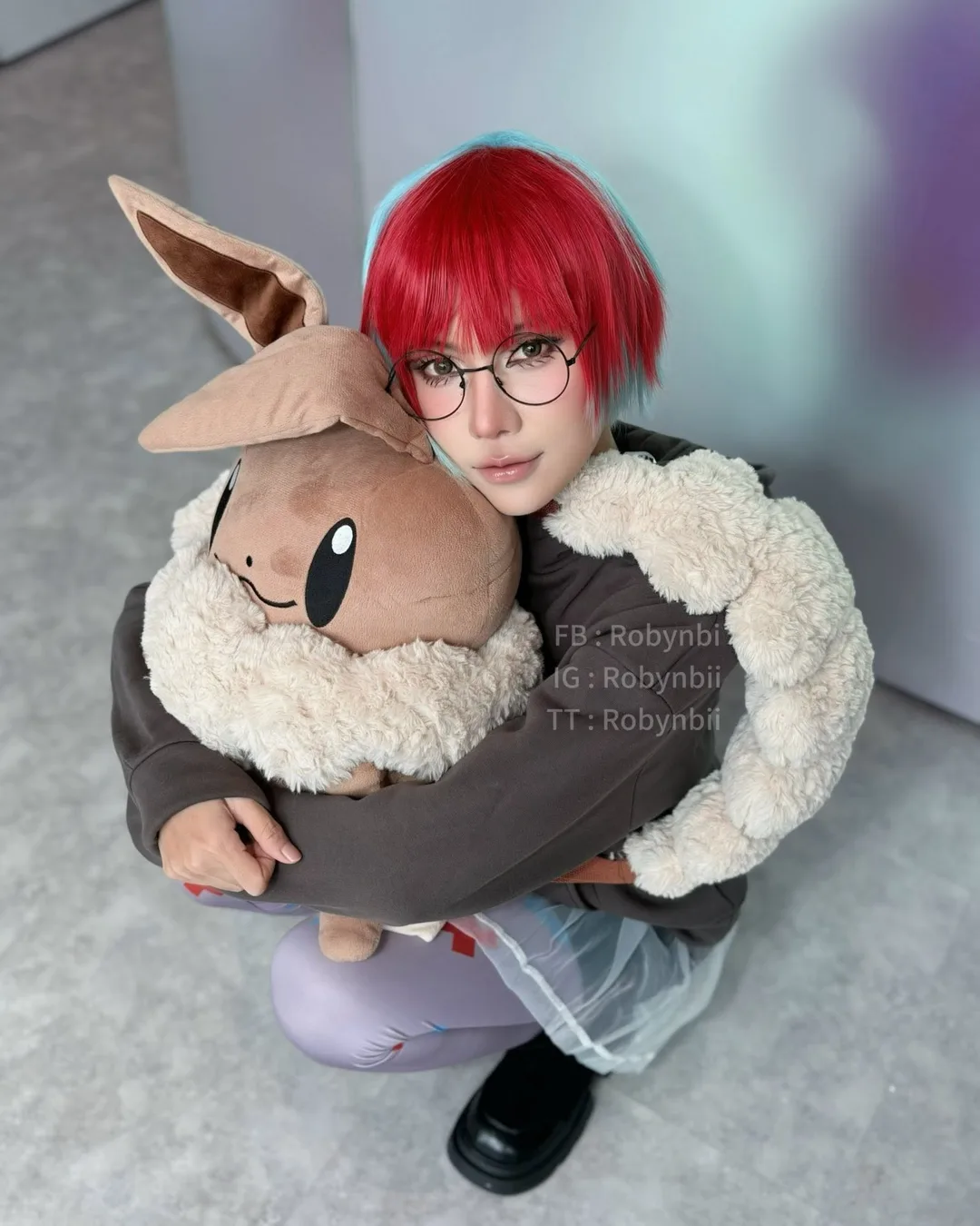 คอสเพลเยอร์แต่งกายเป็น Penny จาก Pokémon นั่งยองๆ ในอาคารพร้อมกอดกระเป๋าเป้ตุ๊กตา Eevee ตัวใหญ่