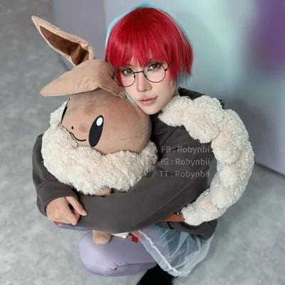 คอสเพลเยอร์แต่งกายเป็น Penny จาก Pokémon นั่งยองๆ ในอาคารพร้อมกอดกระเป๋าเป้ตุ๊กตา Eevee ตัวใหญ่