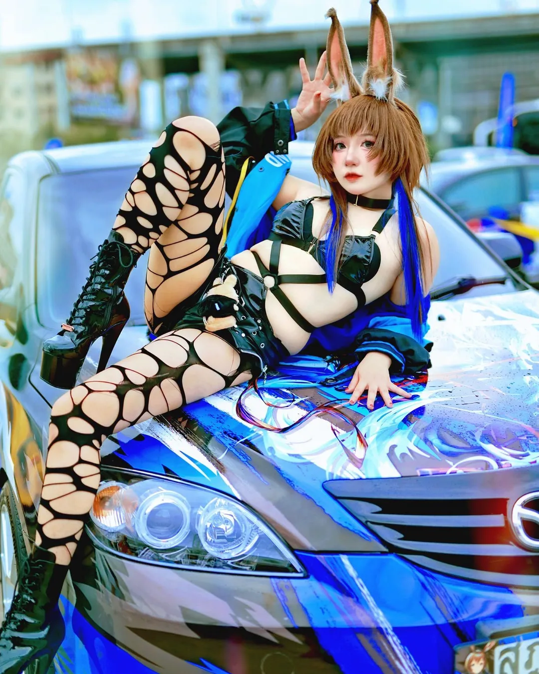 A cosplayer with rabbit ears and ripped tights reclines on the hood of a blue itasha car.