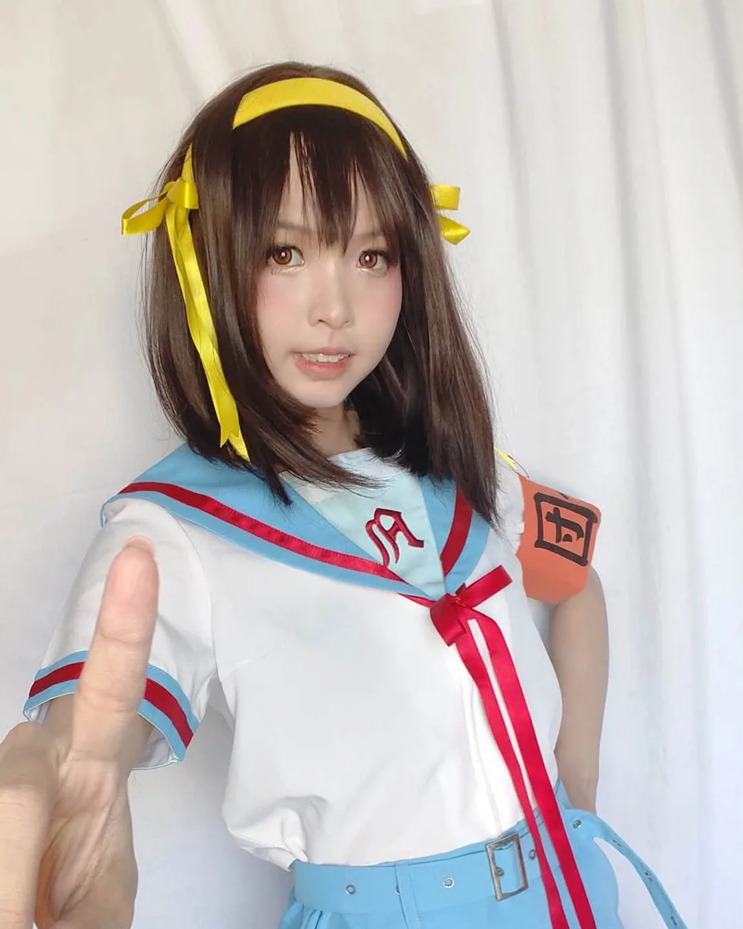 คอสเพลเยอร์แต่งกายเป็น Haruhi Suzumiya ในชุดกะลาสีสีฟ้าขาวชี้นิ้วมาทางกล้องหน้าฉากหลังสีขาว