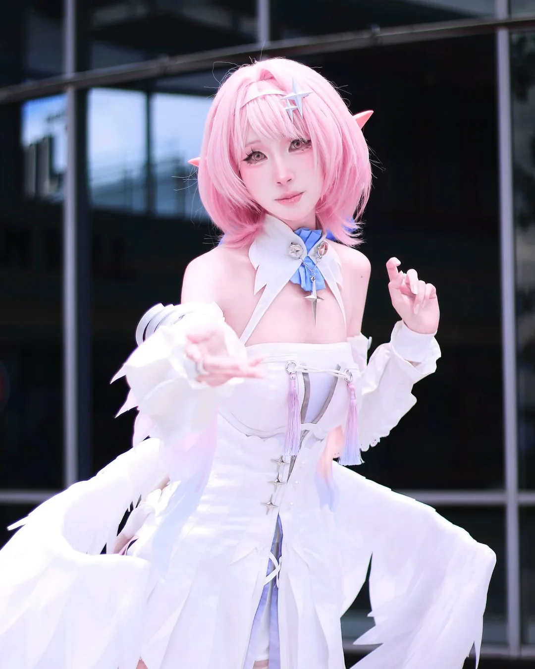 คอสเพลเยอร์ผมสีชมพูติดหูเอลฟ์แต่งชุด Elysia สีขาวติดปีก ยื่นมือข้างหนึ่งมาข้างหน้าขณะโพสท่ากลางแจ้ง