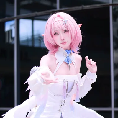คอสเพลเยอร์ผมสีชมพูติดหูเอลฟ์แต่งชุด Elysia สีขาวติดปีก ยื่นมือข้างหนึ่งมาข้างหน้าขณะโพสท่ากลางแจ้ง