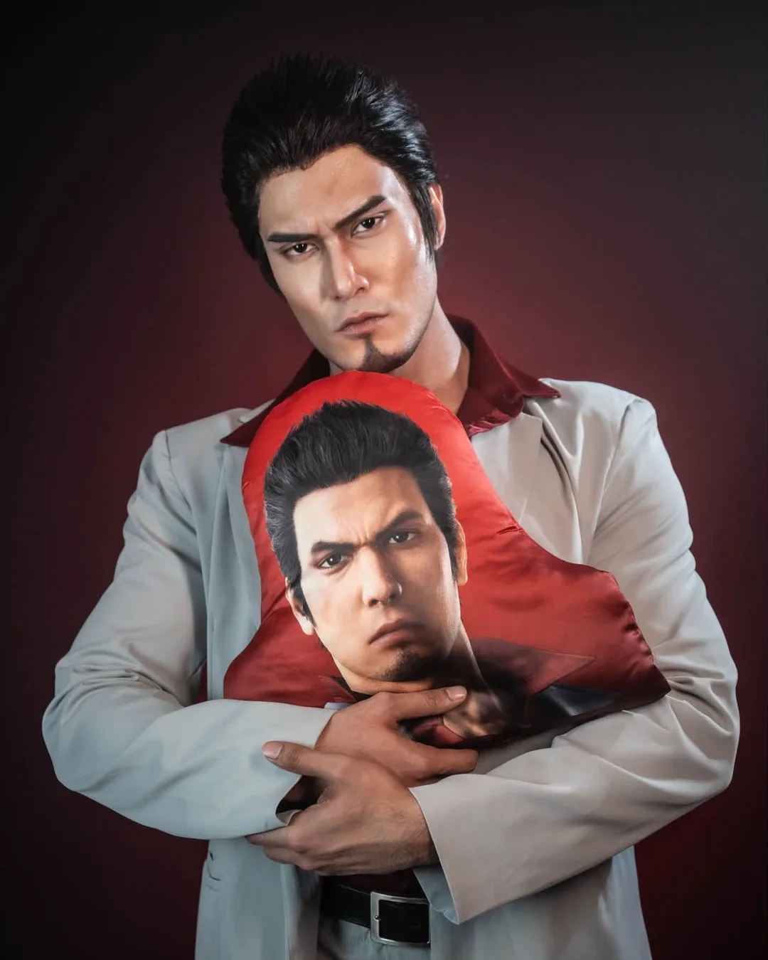 คอสเพลเยอร์ในชุด Kazuma Kiryu จากเกม Yakuza กอดหมอนพิมพ์ลายหน้าของ Kiryu ไว้แนบอกหน้าฉากสตูดิโอสีแดงเข้ม