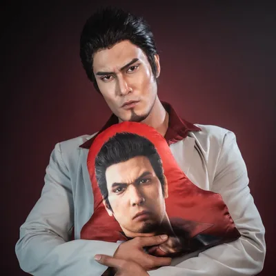 A cosplayer dressed as Kazuma Kiryu from Yakuza hugs a printed pillow of Kiryu's face against a dark red studio backdrop.