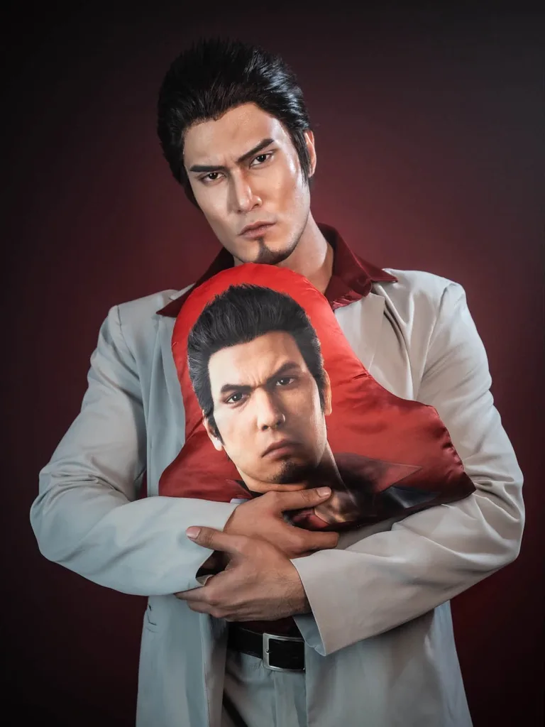 คอสเพลเยอร์ในชุด Kazuma Kiryu จากเกม Yakuza กอดหมอนพิมพ์ลายหน้าของ Kiryu ไว้แนบอกหน้าฉากสตูดิโอสีแดงเข้ม