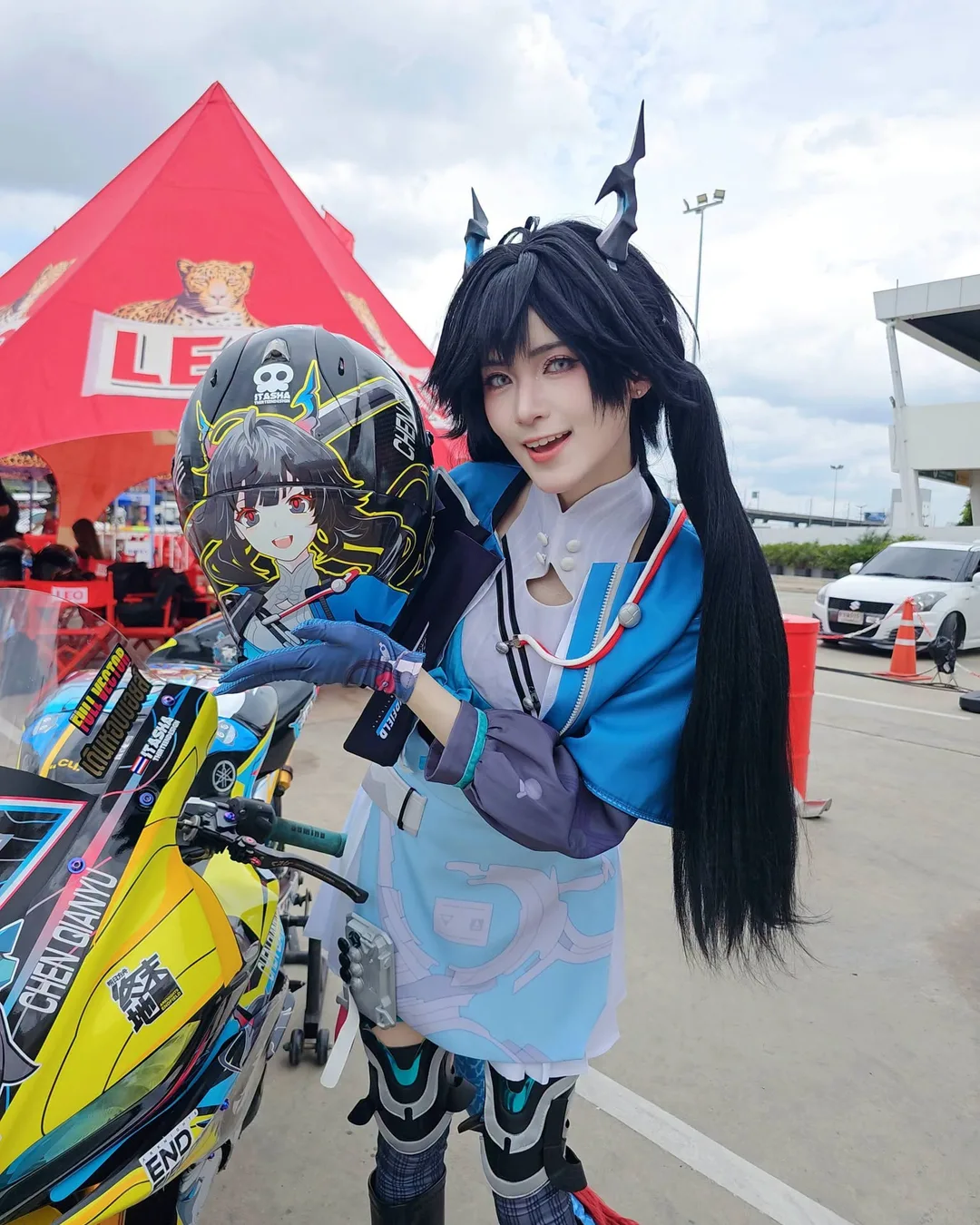 คอสเพลเยอร์ที่มีเขาและมัดผมแกละสีดำยืนโพสท่าข้างมอเตอร์ไซค์ พร้อมถือหมวกกันน็อกลายตัวละครอนิเมะ