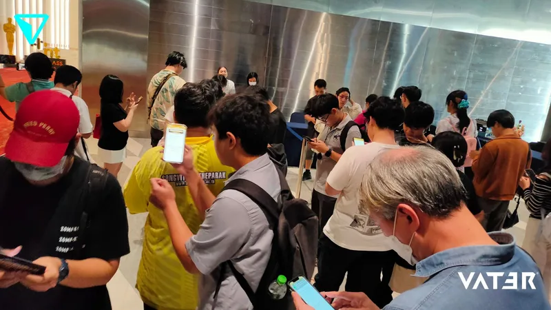 กลุ่มคนกำลังยืนต่อแถวภายในโถงจัดงานพร้อมกับก้มมองสมาร์ตโฟนในมือ