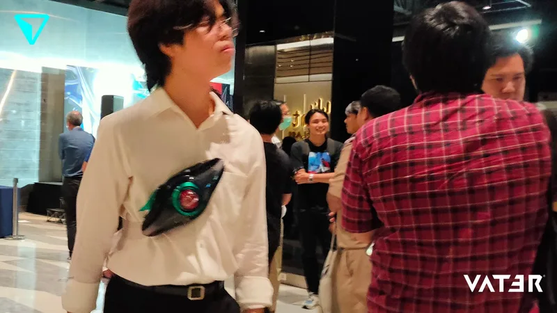 ชายหนุ่มสวมเสื้อเชิ้ตสีขาวเดินอยู่ในงานอีเวนต์ในร่ม โดยสะพายเข็มขัดแปลงร่าง Kamen Rider พาดผ่านหน้าอก
