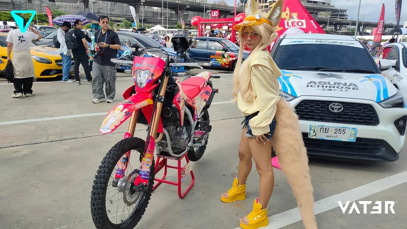 คอสเพลเยอร์สวมหูและหางจิ้งจอกสีบลอนด์ยืนโพสท่ากลางแจ้งระหว่างมอเตอร์ไซค์วิบากสีชมพูและรถยนต์ลายอิตาฉะ
