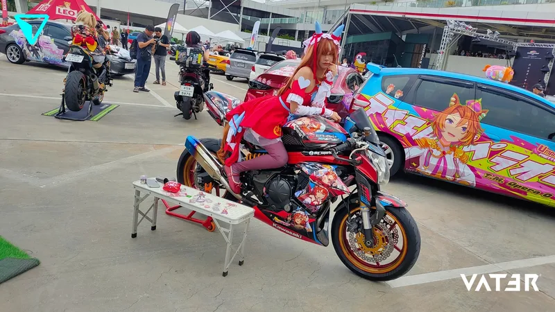 คอสเพลเยอร์ในชุดสีแดงโพสท่าบนมอเตอร์ไซค์ลายอนิเมะในงานจัดแสดงกลางแจ้งข้างรถยนต์ลายอนิเมะ