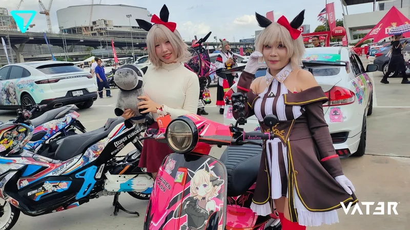 เลเยอร์สองคนในชุดคอสเพลย์อนิเมะยืนโพสท่าด้านหลังสกู๊ตเตอร์ลายอิตาฉะหลากสี โดยมีรถแต่งลายอนิเมะจอดอยู่ด้านหลัง