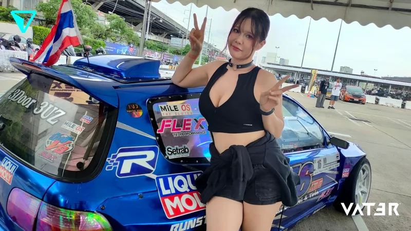 หญิงสาวโพสท่าชูสองนิ้วข้างรถแข่งแต่งซิ่งสีน้ำเงินในงานมอเตอร์สปอร์ตกลางแจ้ง