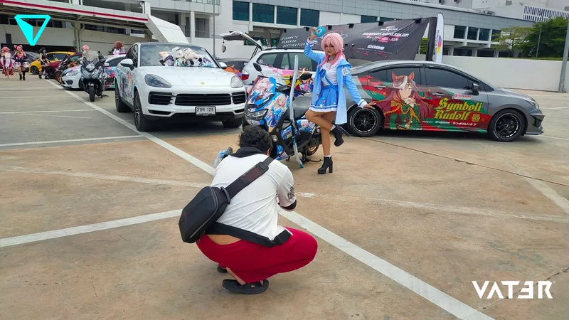 ช่างภาพกำลังนั่งยอง ๆ ถ่ายรูปคอสเพลเยอร์ผมสีชมพูที่กำลังโพสท่าอยู่ข้างรถยนต์และมอเตอร์ไซค์ลายอนิเมะในลานจอดรถ