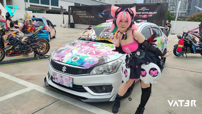 คอสเพลเยอร์ในชุดสีชมพูดำโพสท่าข้างรถยนต์ Suzuki ลายการ์ตูนอนิเมะในงานแสดงรถยนต์กลางแจ้ง