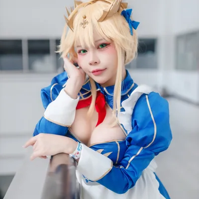 คอสเพลเยอร์ในชุดเมดสีน้ำเงินและขาวสไตล์ Artoria Pendragon เท้าแขนกับราวระเบียงพร้อมมองกล้องในอาคาร