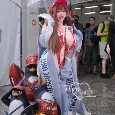 A cosplayer in a red-and-white swimsuit stands under a clear umbrella next to lifeguard penguin statues at an outdoor event.
