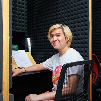 คนผมบลอนด์ยิ้มขณะนั่งอยู่ที่โต๊ะพร้อมเอกสารบทพากย์ในห้องอัดเสียงที่บุด้วยโฟมซับเสียง