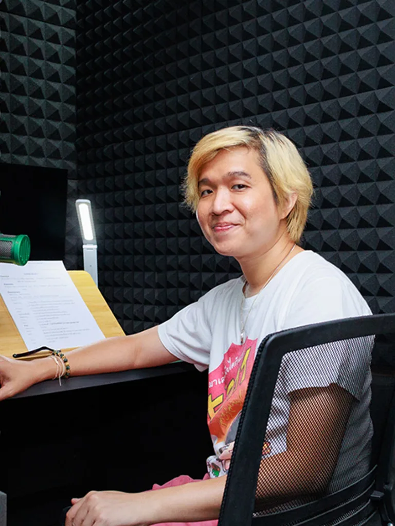 คนผมบลอนด์ยิ้มขณะนั่งอยู่ที่โต๊ะพร้อมเอกสารบทพากย์ในห้องอัดเสียงที่บุด้วยโฟมซับเสียง