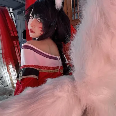 A cosplayer dressed as Ahri from League of Legends looks over her shoulder, surrounded by large, fluffy white fox tails.