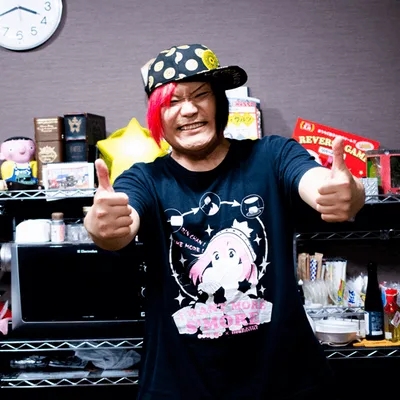 A smiling man with pink hair highlights and a cap gives two thumbs up in front of shelves filled with anime and gaming merchandise.