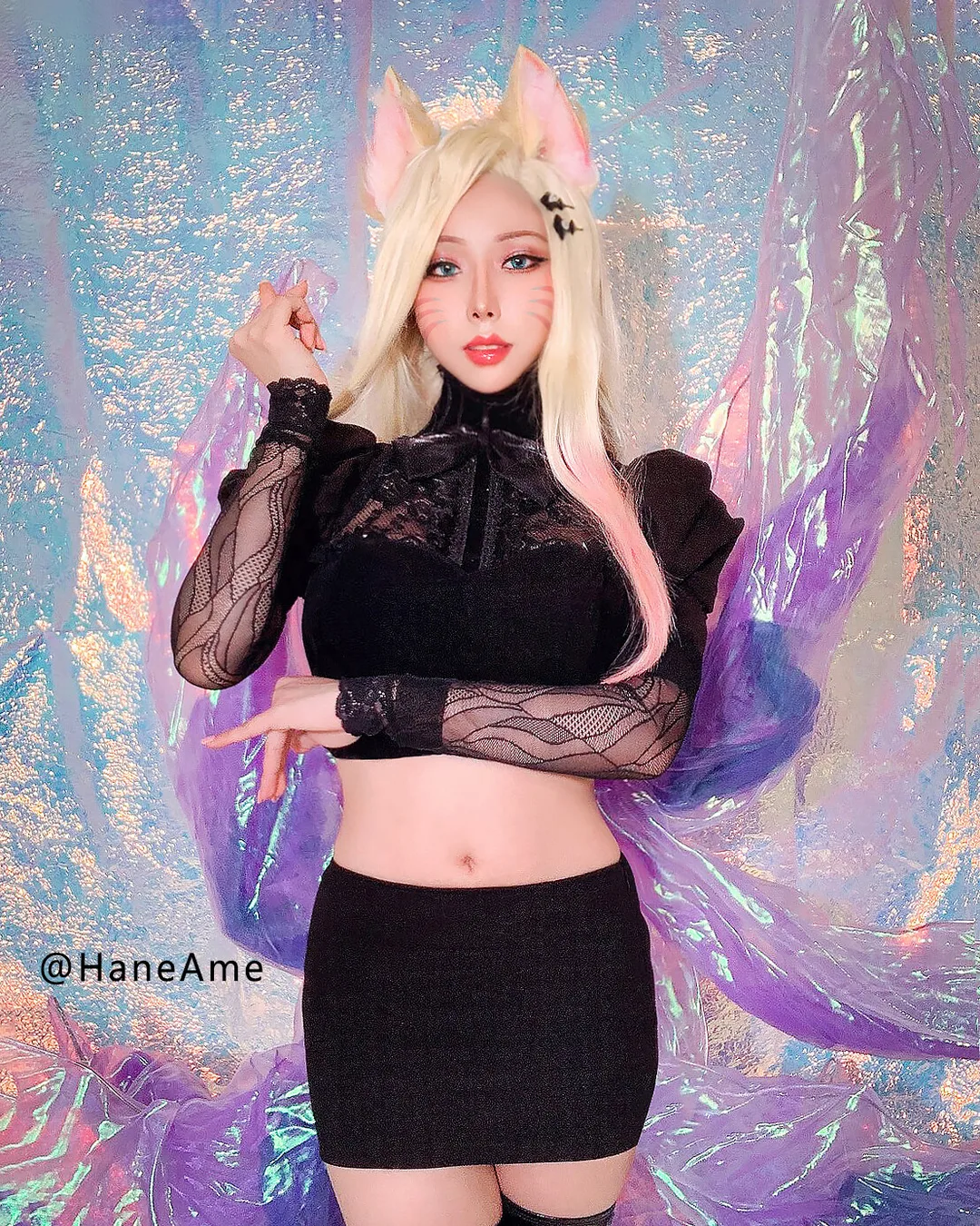 A cosplayer dressed as Ahri from League of Legends poses in a black outfit, blonde wig, and fox ears before an iridescent backdrop.