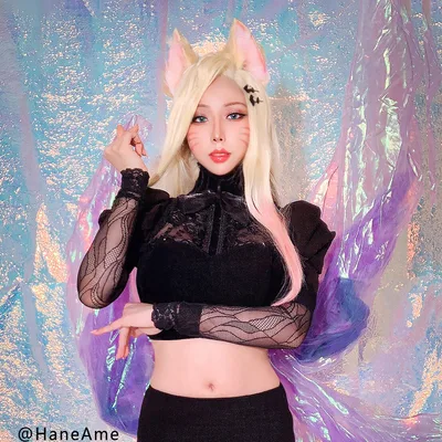 A cosplayer dressed as Ahri from League of Legends poses in a black outfit, blonde wig, and fox ears before an iridescent backdrop.